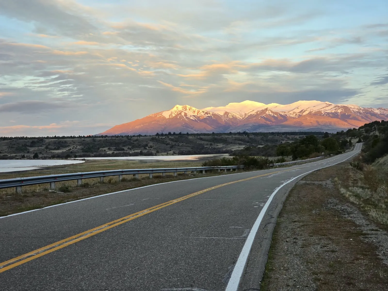Ruta junto a un lago con la cordillera nevada iluminada al atardecer